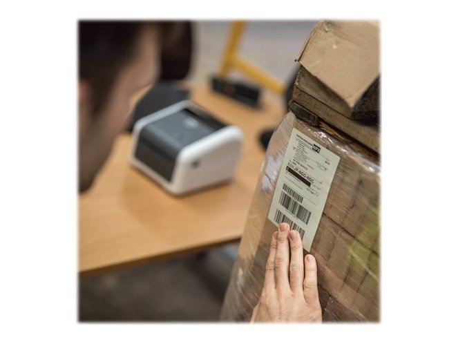 Copy electronic vam predstavlja Printer BROTHER TD-4420DN Label direct thermal, LAN [TD4420DNXX1]. Na fotografiji je kutija na kojoj je zaljepljen naljepnica. U pozdani je stolu je printer. Pored kutije je muškarac koji drži ruku na kutiji