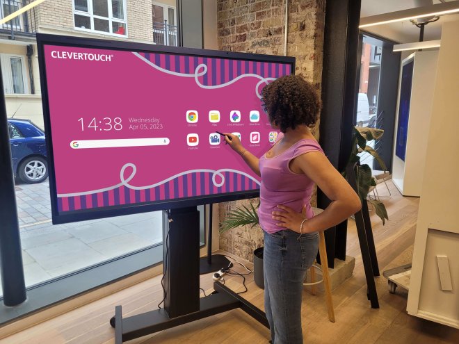 Copy electronic vam predstavlja Interaktivni ekran Clevertouch 65" Impact LUX, DLED 4K, Android 13, 8GB, 128GB. Na fotografiji je ured u kojem je zenska osoba koja stoji ispred ekrana. Ekran je postavljen na crno postolje. Na ekrau je pozadina crvene boje.